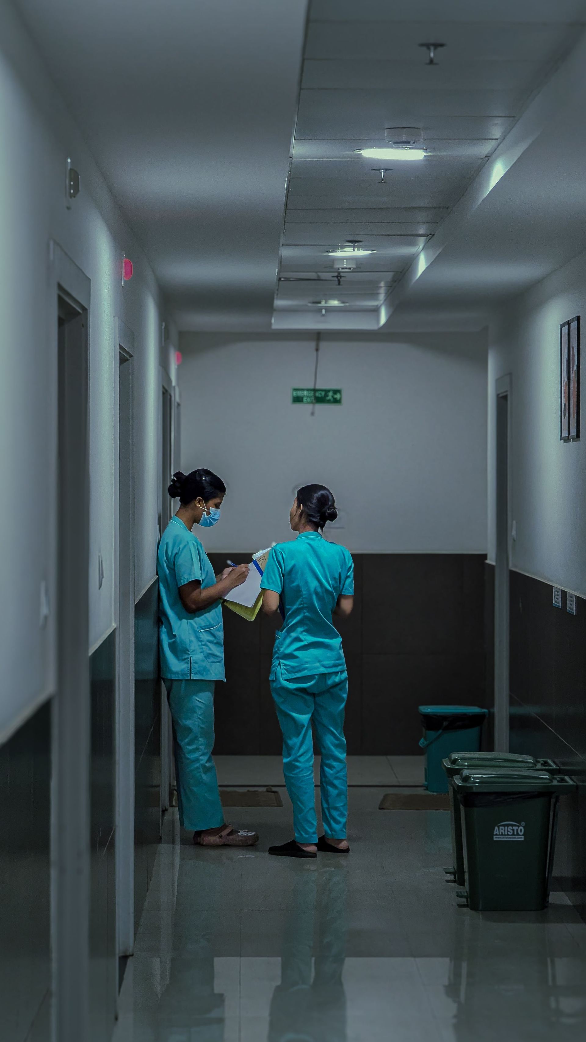 Duas profissionais de enfermagem de uniforme azul-esverdeado conversam num corredor de hospital, uma delas de máscara, conferindo papéis numa prancheta.