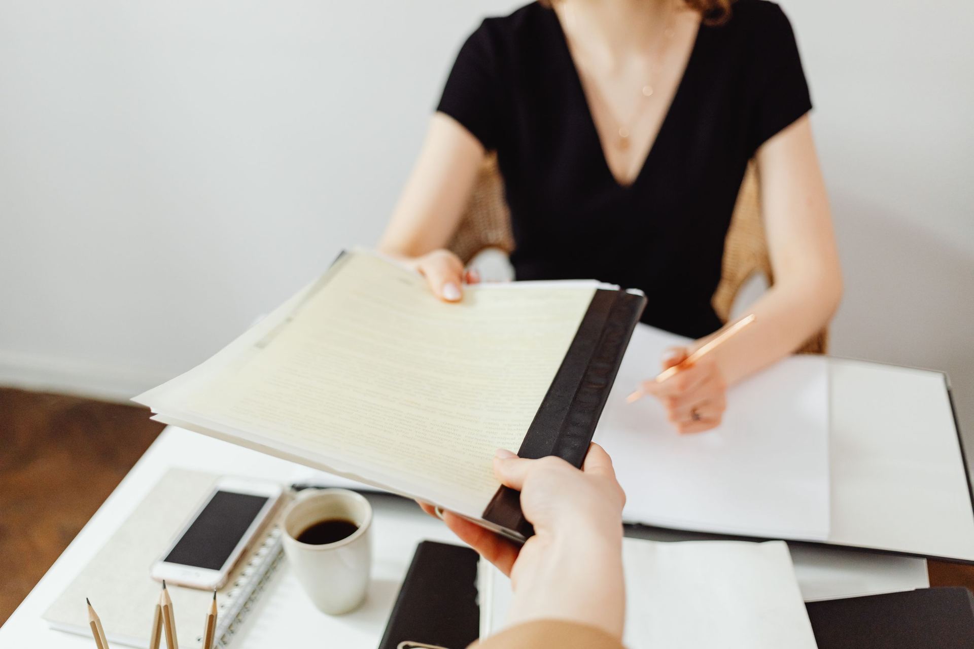 Uma pessoa entrega a outra, por cima da mesa, um documento encadernado com várias páginas, ao lado de uma xícara de café e de um bloco de anotações.