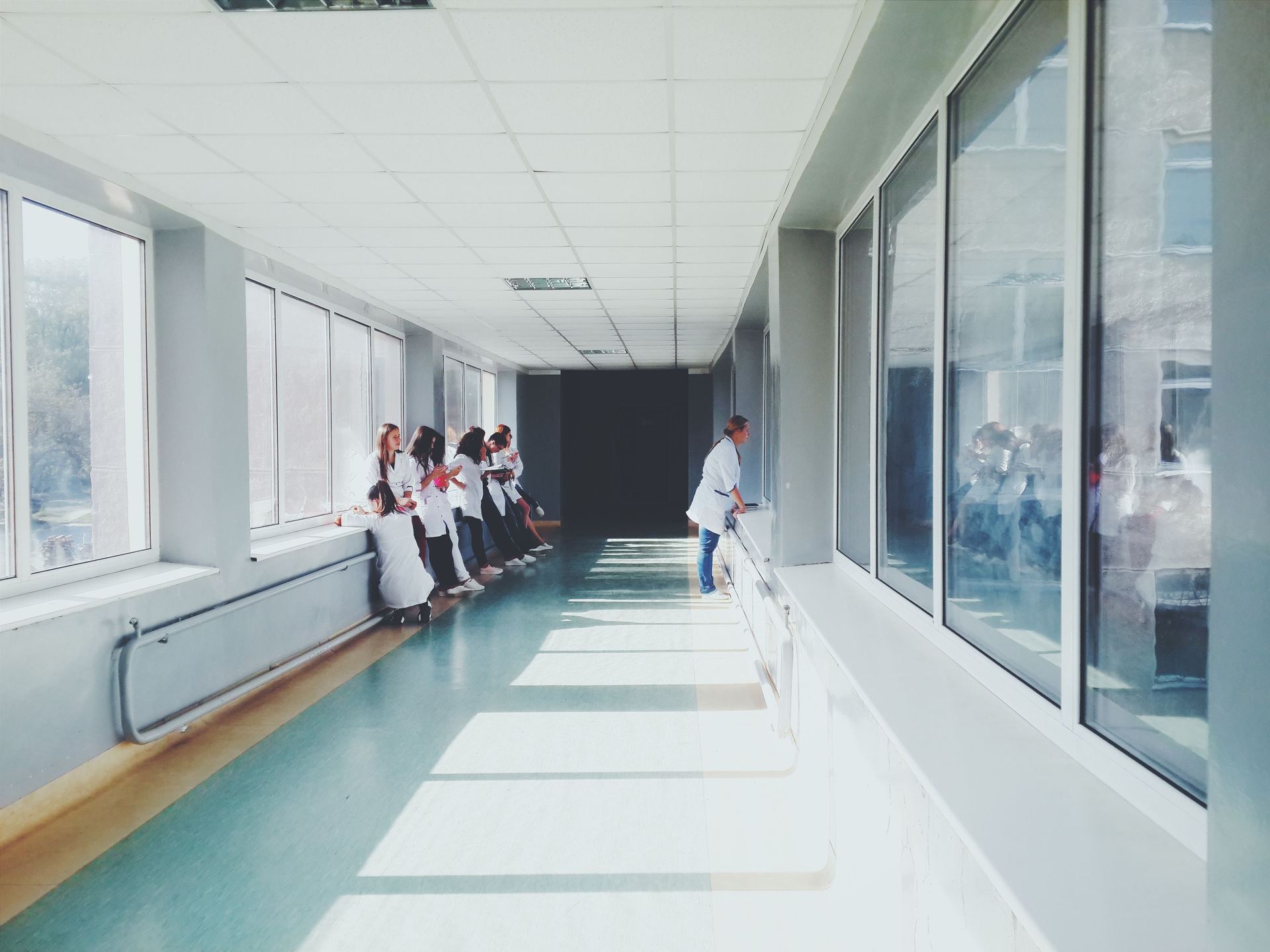 Corredor de hospital iluminado, com profissionais de branco encostados na janela